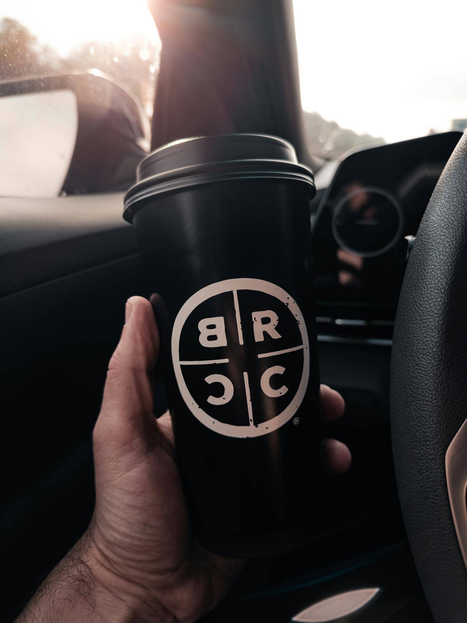 A hand holding a branded coffee cup inside a car, bathed in warm morning light.