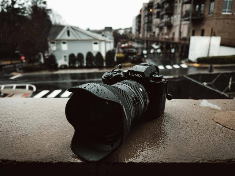 A Lumix camera sits on a wet balcony railing on a rainy day in the city.