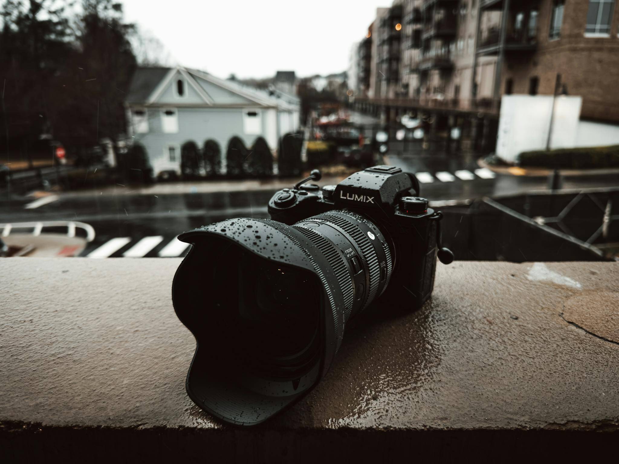 A Lumix camera sits on a wet balcony railing on a rainy day in the city.
