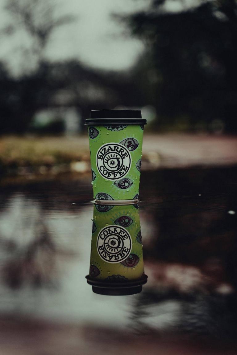 A unique coffee cup with eye designs reflected in a calm puddle outdoors.